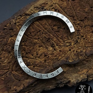 Könnte beinhalten: Ein silbernes Armband mit einem runden Design, das mit nordischen Runen verziert ist, die um das Band graviert sind.