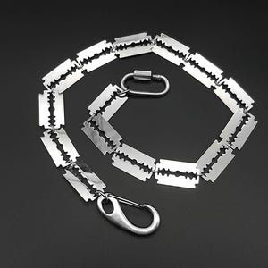 Könnte beinhalten: Eine silberfarbene Halskette mit einer Reihe von Rasierklingen-Anhängern. Die Kette ist mit einem Karabinerverschluss und einem Schraubverschluss verbunden. Der Hintergrund ist einfarbig schwarz.