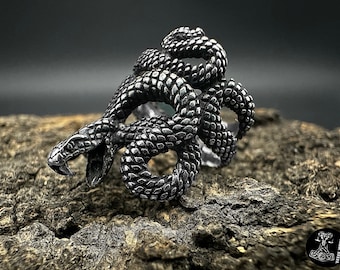May include: A silver ring with a detailed design of three intertwined snakes. The snakes are coiled together and have a textured, scaly appearance.