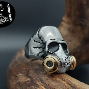Könnte beinhalten: Ein silberner Ring mit einem Totenkopf, der eine Gasmaske trägt. Die Gasmaske hat goldene Akzente. Der Ring liegt auf einer Holzoberfläche.
