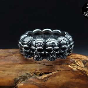 Könnte beinhalten: Ein silberner Ring mit einem Design aus mehreren Schädeln um das Band. Der Ring liegt auf einem Stück Holz.