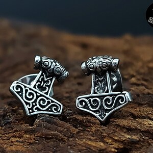 Puede incluir: Un par de pendientes de botón de plata con un diseño detallado de martillo Mjolnir con intrincados patrones de espiral.