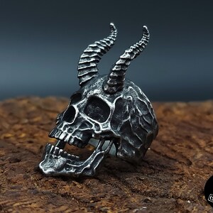 Könnte beinhalten: Ein silberner Totenkopfring mit Hörnern, der Totenkopf ist mit einer strukturierten Oberfläche detailliert und hat einen offenen Mund.
