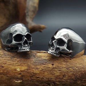 Puede incluir: Dos anillos de calavera plateados sobre una superficie de madera. Los anillos están hechos de acero inoxidable y tienen un diseño de calavera detallado.