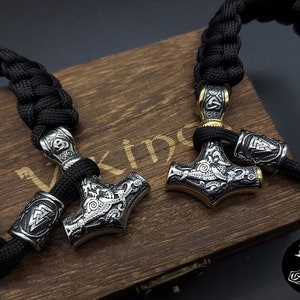 Könnte beinhalten: Zwei schwarze Paracord-Armbänder mit silbernen und goldenen Mjolnir-Anhängern. Die Anhänger sind mit aufwendigen Gravuren verziert. Die Armbänder liegen auf einer Holzoberfläche mit der eingravierten Aufschrift "Viking".