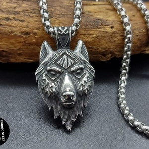 Può includere: Una collana con un ciondolo a forma di testa di lupo in argento, con un design dettagliato e una catena. Il ciondolo presenta il viso di un lupo con un motivo geometrico sulla fronte. La catena è una catena a scatola d'argento.