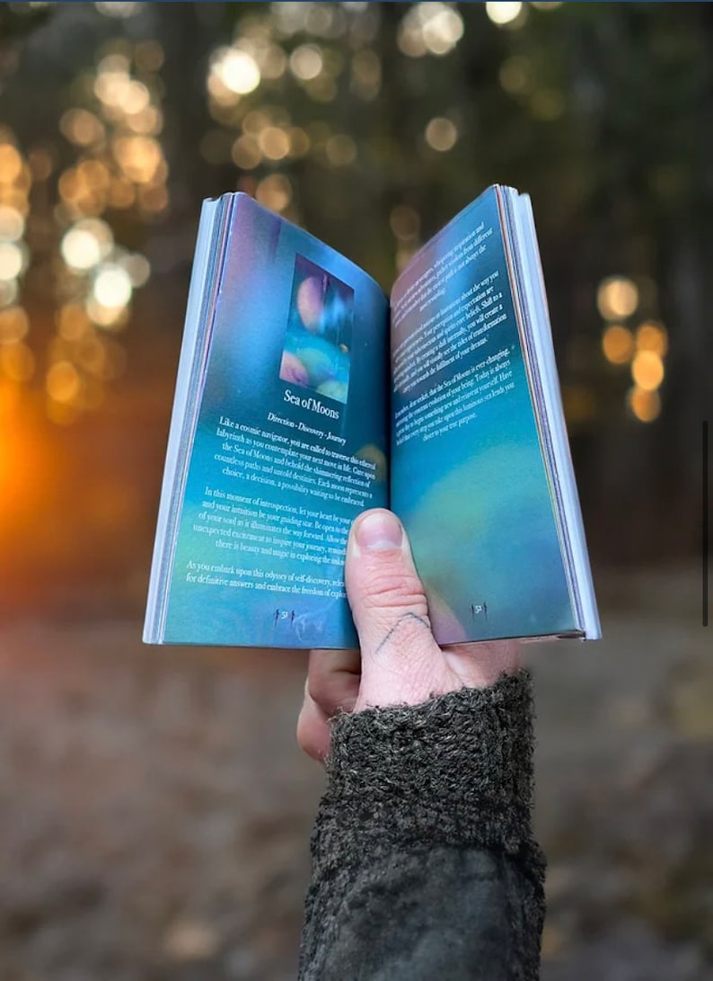 K&ouml;nnte beinhalten: Ein blaues und violettes Buch mit dem Titel "Sea of Moons" und dem Text "Direction: Discovery Journey" auf dem Cover. Das Buch ist auf einer Seite mit einer blauen und violetten Illustration eines Mondes und Sternen ge&ouml;ffnet.