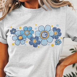 May include: A gray t-shirt with a blue and yellow floral design. The flowers have a yellow center and the letters "KC" are in the center of the largest flower.