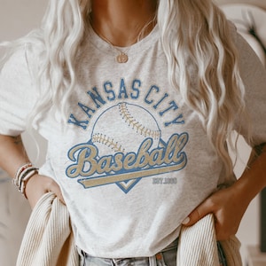 May include: A light gray t-shirt featuring a baseball design. The design includes the words "Kansas City Baseball" in blue and gold, with a baseball graphic in the center. The text "EST. 1969" is also included.