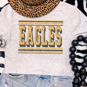 May include: A light gray crop top with the word "EAGLES" in gold and black lettering. The shirt has a vintage, distressed look. A leopard print hat is partially visible at the top, and a black beaded necklace is on the right.