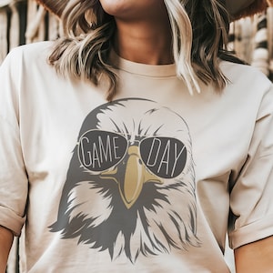 May include: A beige t-shirt with a graphic of an eagle wearing sunglasses and the text "GAME DAY".