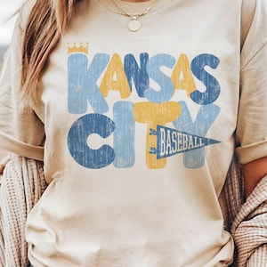 Pu&ograve; includere: Una maglietta beige con un design grafico in stile vintage che dice "Kansas City Baseball" in lettere blu, gialle e bianche.