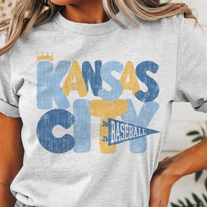 Pu&ograve; includere: Una maglietta bianca con una grafica in stile vintage che dice "Kansas City" in lettere blu e gialle con una corona sopra la "K" e un pennant che dice "Baseball" sotto la "Y".