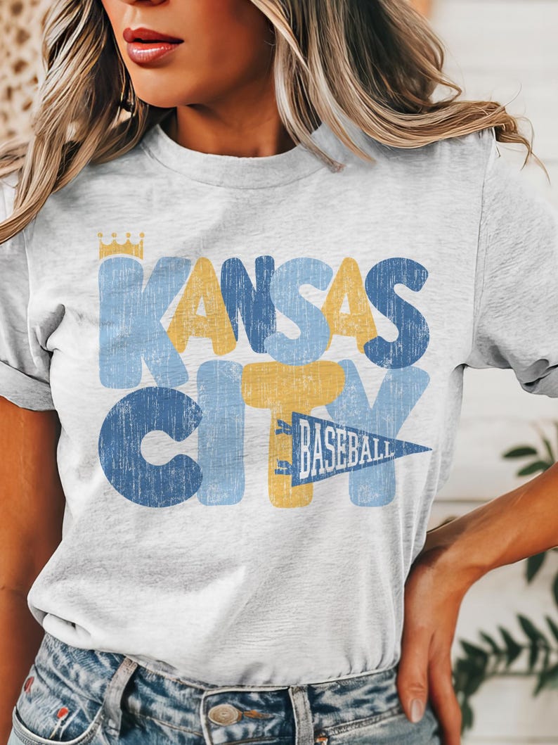 Pu&ograve; includere: Una maglietta bianca con una grafica in stile vintage che dice "Kansas City" in lettere blu e gialle con una corona sopra la "K" e un pennant che dice "Baseball" sotto la "Y".