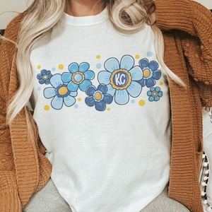 May include: A white t-shirt with a blue and yellow floral design. The flowers have yellow centers and the largest flower has the letters "KC" in the center.