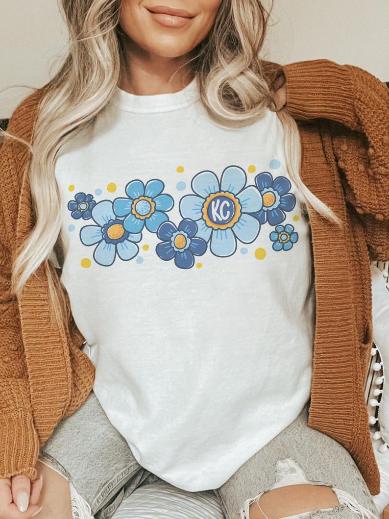 May include: A white t-shirt with a blue and yellow floral design. The flowers have yellow centers and the largest flower has the letters "KC" in the center.