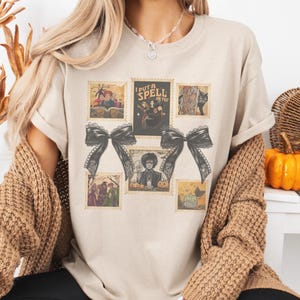 May include: Beige t-shirt featuring a collage of vintage-style postage stamps with Halloween-themed illustrations. The stamps include images of witches, black cats, and the text "I PUT A SPELL ON YOU". Two large black bows are also featured.