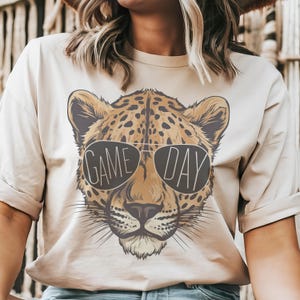 May include: A beige t-shirt with a cheetah print graphic wearing sunglasses that say "GAME DAY".