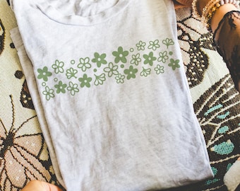 Könnte beinhalten: Hellgraues T-Shirt mit einem grünen Blumenmuster. Die Blumen sind in einer horizontalen Reihe angeordnet. Das T-Shirt ist gefaltet und wird von Händen gehalten.