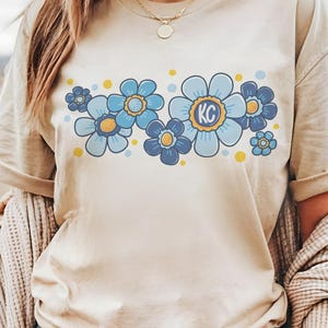 May include: A beige t-shirt with a graphic of blue and yellow flowers. The center flower has the letters "KC" in the middle.
