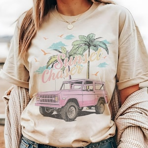 May include: A beige t-shirt with a pink vintage jeep graphic and the text "Sunset Chaser" in a colorful font. Palm trees and clouds are in the background.