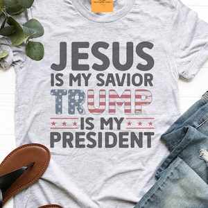 May include: Gray t-shirt with the text "JESUS IS MY SAVIOR TRUMP IS MY PRESIDENT" in black. The word "TRUMP" is in red, white, and blue with stars.