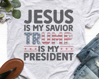 May include: Gray t-shirt with the text "JESUS IS MY SAVIOR TRUMP IS MY PRESIDENT" in black. The word "TRUMP" is in red, white, and blue with stars.