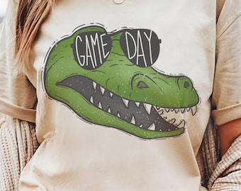 May include: A beige t-shirt with a green alligator wearing sunglasses and the text "GAME DAY" printed on the front.