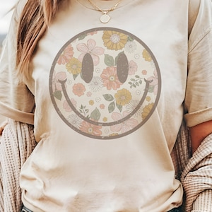 May include: Beige t-shirt featuring a floral smiley face design. The smiley face is filled with pink, yellow, and white flowers. The shirt has short sleeves and a crew neck. The Marigold Lane logo is in the upper left corner.