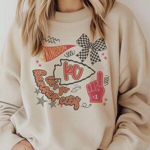 May include: A beige sweatshirt with a graphic design featuring the text "Kansas City" and "Kingdom". The design includes a KC arrowhead, a checkered bow, a foam finger, and stars. The sweatshirt is a neutral color.
