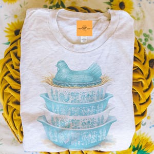 May include: A light gray t-shirt featuring a blue hen on a nest design above three stacked turquoise Pyrex bowls. The bowls have a white and turquoise pattern. The shirt has a small orange tag that says "marigold lane."
