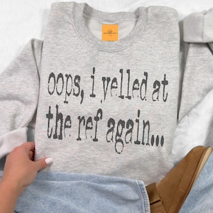 May include: Gray sweatshirt with the text "oops, i yelled at the ref again..." printed on the front.