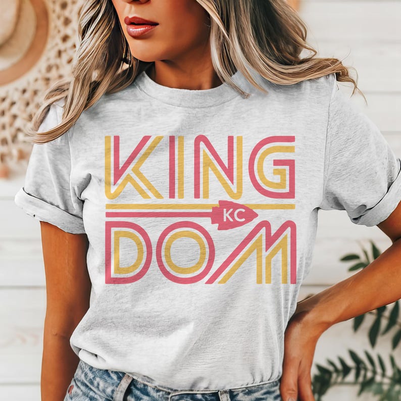 May include: A light gray t-shirt with the word "KINGDOM" in yellow and pink lettering. The word "KC" is in the center of the shirt, with an arrow pointing to the right. The shirt is made of a soft material.