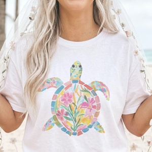 May include: White t-shirt featuring a colorful watercolor turtle design. The turtle's shell is filled with pink, yellow, and blue flowers and green leaves. The shirt is a classic crew neck style.