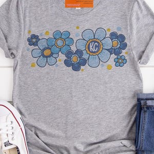 May include: Gray t-shirt with a blue and gold floral design. The flowers have a retro style and the center flower has the letters "KC" in the middle.