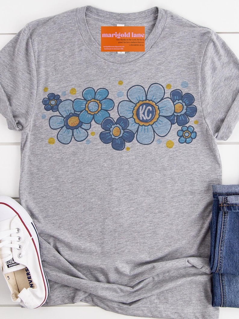 May include: Gray t-shirt with a blue and gold floral design. The flowers have a retro style and the center flower has the letters "KC" in the middle.