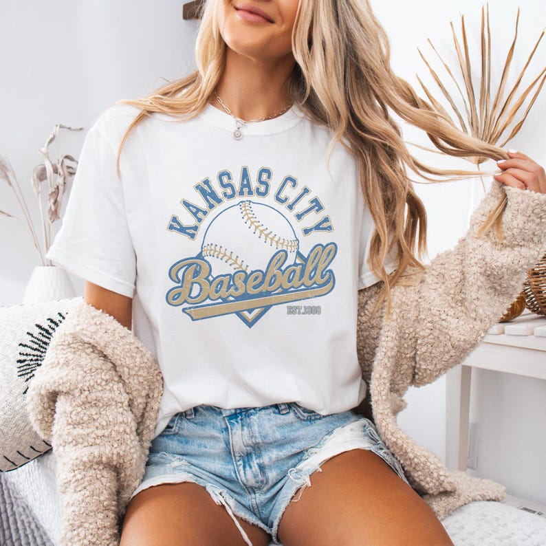 May include: White t-shirt with a baseball graphic. The design features the text "Kansas City Baseball" in blue and gold, with a baseball illustration. The shirt has a crew neck and short sleeves. The text "EST.1889" is also included.
