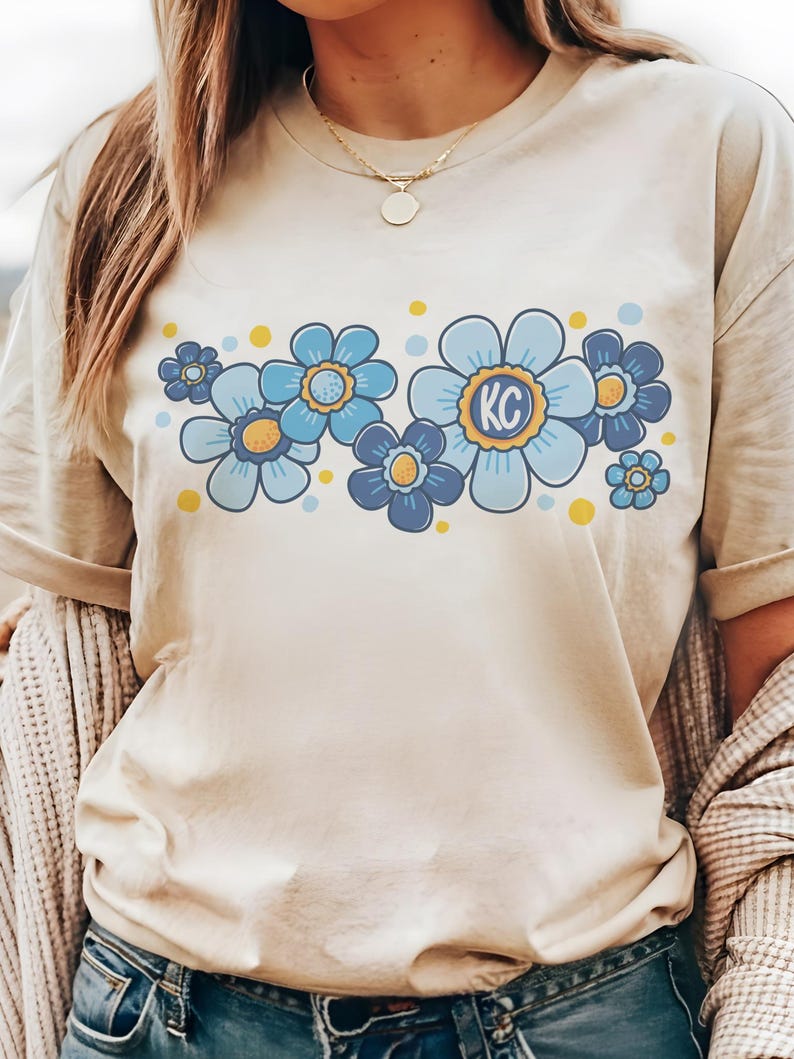 May include: A beige t-shirt with a graphic of blue and yellow flowers. The center flower has the letters "KC" in the middle.