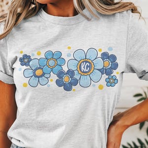 May include: A gray t-shirt with a blue and yellow floral design. The flowers have a yellow center and the letters "KC" are in the center of the largest flower.