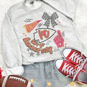 May include: A gray sweatshirt with a graphic that says "Let's go Kansas City" with a KC outline, a bow, and a foam finger. The image also includes red sneakers, denim shorts, and a football.