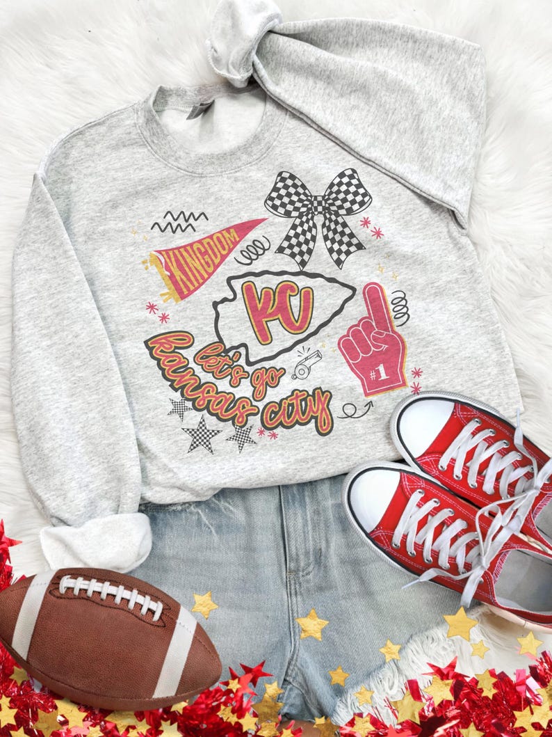 May include: A gray sweatshirt with a graphic that says "Let's go Kansas City" with a KC outline, a bow, and a foam finger. The image also includes red sneakers, denim shorts, and a football.