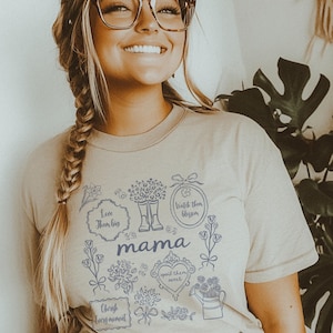 May include: Beige t-shirt with the word "mama" in the center, surrounded by blue floral illustrations and phrases like "Love them big" and "Cherish every moment." The shirt has a crew neck and short sleeves.