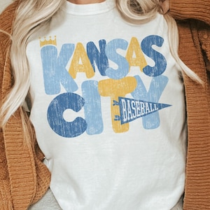 Pu&ograve; includere: Maglietta bianca con una grafica in stile vintage che dice "Kansas City" in lettere blu, gialle e bianche con un pennant di baseball che dice "Baseball" in lettere gialle su un pennant blu.