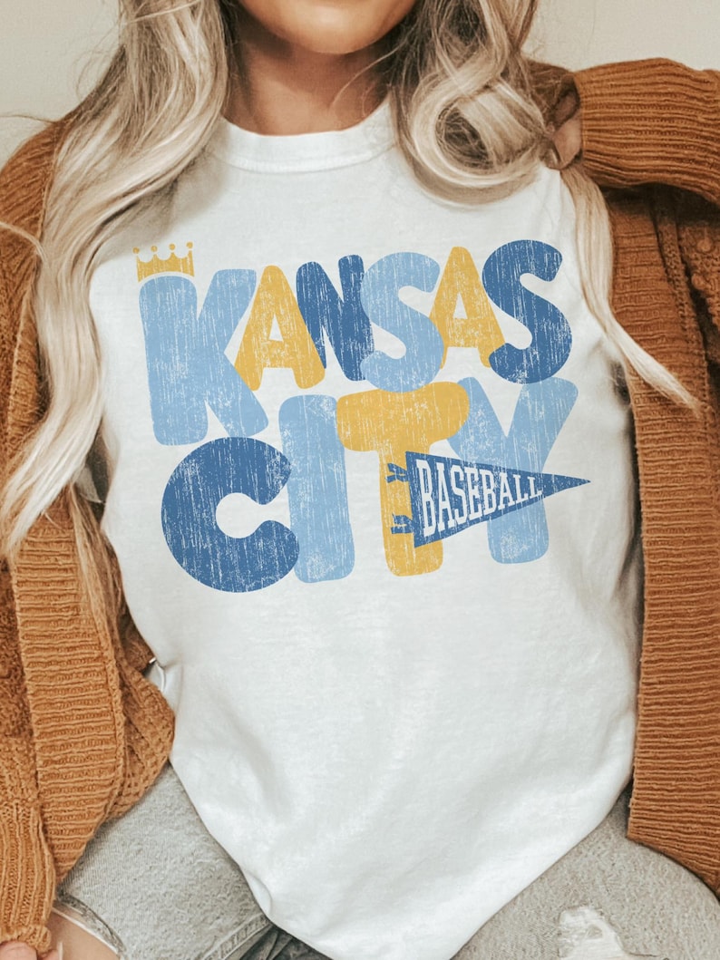 Pu&ograve; includere: Maglietta bianca con una grafica in stile vintage che dice "Kansas City" in lettere blu, gialle e bianche con un pennant di baseball che dice "Baseball" in lettere gialle su un pennant blu.