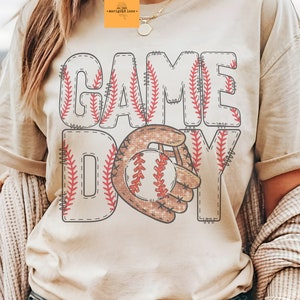 以下が含まれることがあります： 野球をテーマにしたグラフィックが特徴のクリーム色のTシャツ。「GAME DAY」の文字がステッチフォントで表示され、野球とグローブのグラフィックが描かれています。ゆったりとしたフィット感のシャツです。