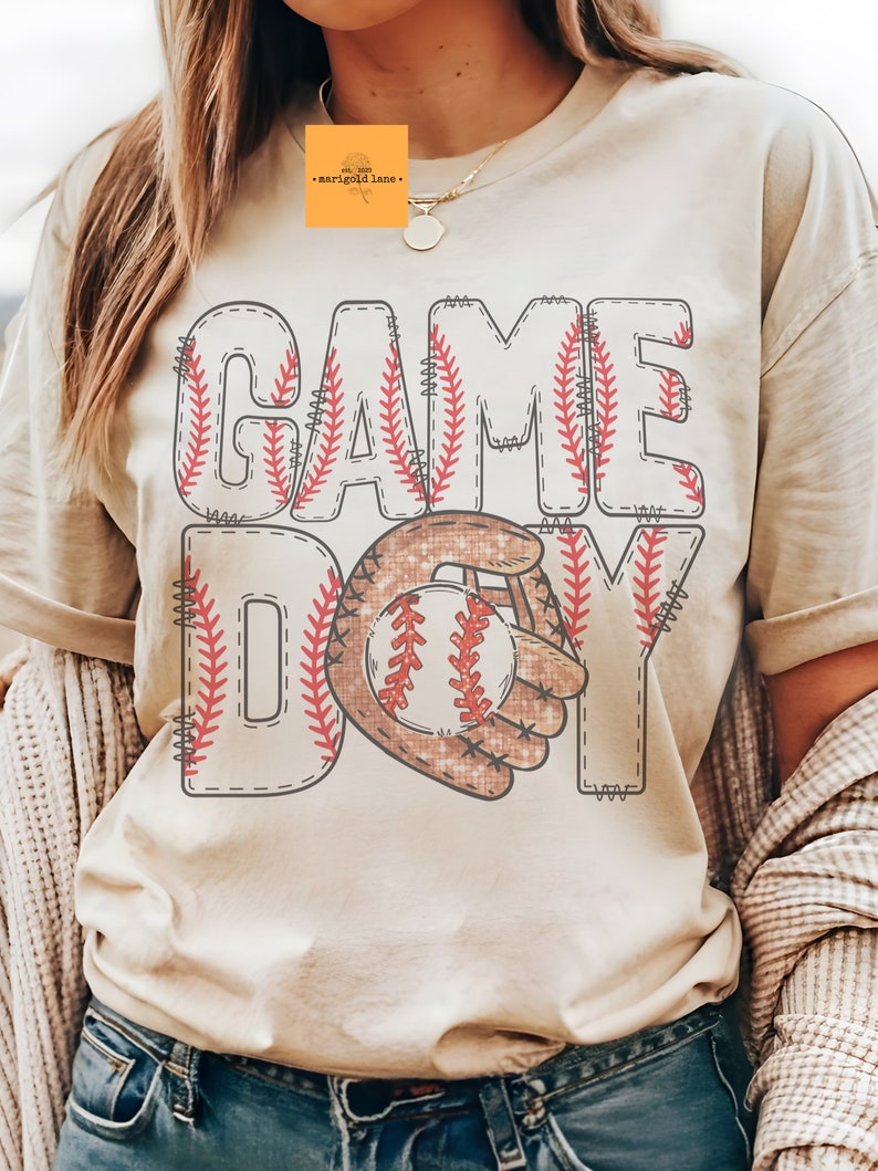 以下が含まれることがあります： 野球をテーマにしたグラフィックが特徴のクリーム色のTシャツ。「GAME DAY」の文字がステッチフォントで表示され、野球とグローブのグラフィックが描かれています。ゆったりとしたフィット感のシャツです。