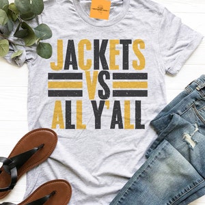 May include: A gray t-shirt with the text "Jackets vs All Y'all" in black and gold lettering. The shirt has a distressed look, with a faded background and a slightly worn appearance.