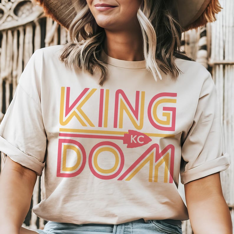 May include: Beige t-shirt with the words "KINGDOM KC" in yellow and pink. The word "KING" is above "DOM", with an arrow pointing to the letters "KC". The shirt has rolled-up sleeves.