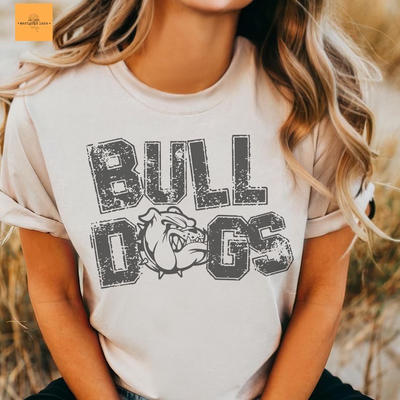 Puede incluir: Camiseta beige claro con la inscripci&oacute;n "BULL DOGS" en letras grises desgastadas. El dise&ntilde;o incluye una ilustraci&oacute;n de bulldog. La camiseta tiene mangas cortas y cuello redondo.