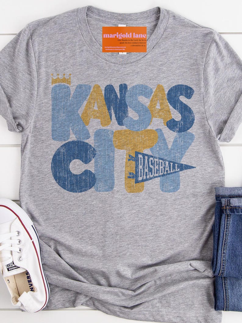 Pu&ograve; includere: Maglietta grigia con una grafica in stile vintage che dice "Kansas City Baseball" in lettere blu, gialle e bianche con una corona sopra la parola "Kansas".